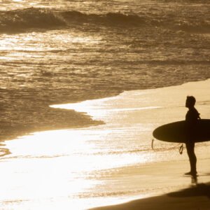 Silhouetted, isolated surfer standing on golden sand before sunwashed waves