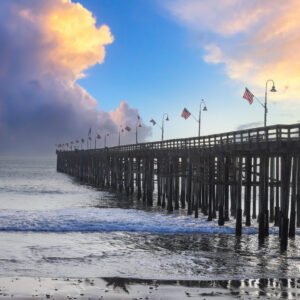 a long brown wooden pier with American flags flying from curved light posts surrounded by vast blue ocean water with blue sky and clouds at Ventura Pier in Ventura California USA